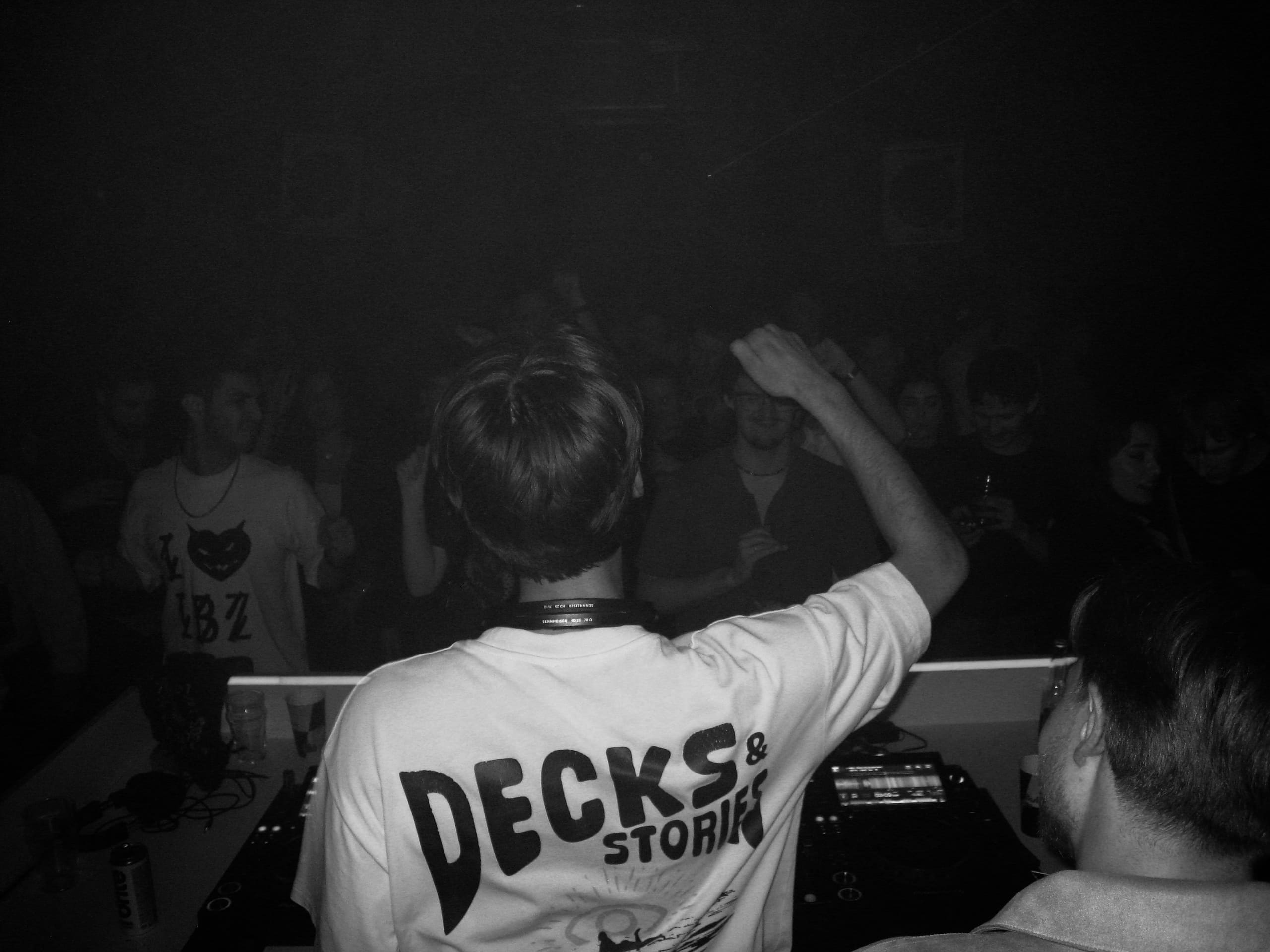 Aksendo from behind at the decks, fist raised, Decks & Stories shirt — live press portrait.